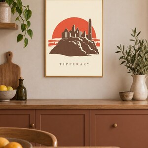 May include: A rectangular wall art print featuring a stylized illustration of the Rock of Cashel in Tipperary, Ireland. The design displays a dark brown castle silhouette against a large red sun, set on a cream background with the text TIPPERARY.