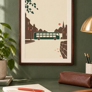 May include: A framed art print featuring a stylized illustration of a dark green double-decker bus driving through a town street. The scene includes brown silhouettes of buildings, a small red sun, a street lamp, and green tree leaves in the corner.