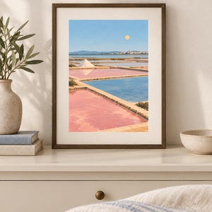 May include: A framed art print depicting a landscape of salt pans with rectangular pools of pink and blue water, a white salt mound, distant hills, and a pale yellow sun in a clear blue sky, displayed in a dark brown wooden frame on a white wall.
