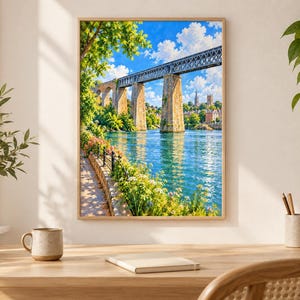 May include: A framed wall art print depicting a stone viaduct bridge spanning a blue river. The scene includes a sunlit riverside path with green foliage, blooming flowers, and a distant town skyline under a bright blue sky with white clouds.