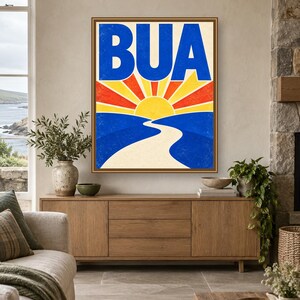 May include: A framed wall art print featuring a stylized landscape with a bright yellow sun rising over blue hills. A winding white path leads toward the horizon. The bold blue text BUA is centered at the top against a cream background with red and yellow sunburst rays.