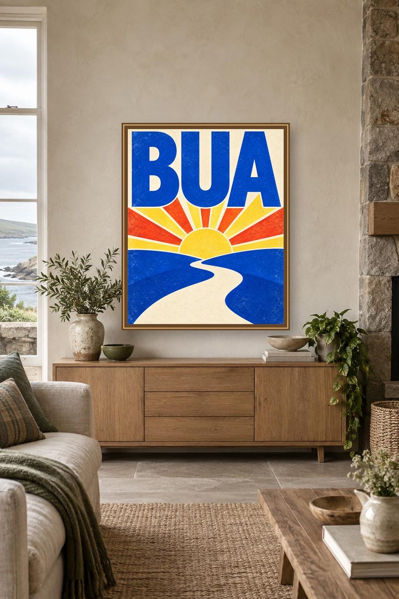 May include: A framed wall art print featuring a stylized landscape with a bright yellow sun rising over blue hills. A winding white path leads toward the horizon. The bold blue text BUA is centered at the top against a cream background with red and yellow sunburst rays.