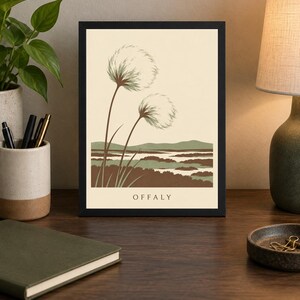 May include: A framed art print featuring a cream background with two fluffy white seed heads on long brown stems in the foreground. Below, a stylized landscape of green and brown marshland is depicted. The word OFFALY is printed in black at the bottom.