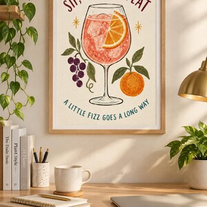 Op de afbeelding: Een ingelijste kunstprint met een cocktail in een glas op voet met een schijfje sinaasappel en ijsblokjes. De tekst luidt bovenaan SIP, SMILE, REPEAT en onderaan A LITTLE FIZZ GOES A LONG WAY, met illustraties van druiven en een sinaasappel.