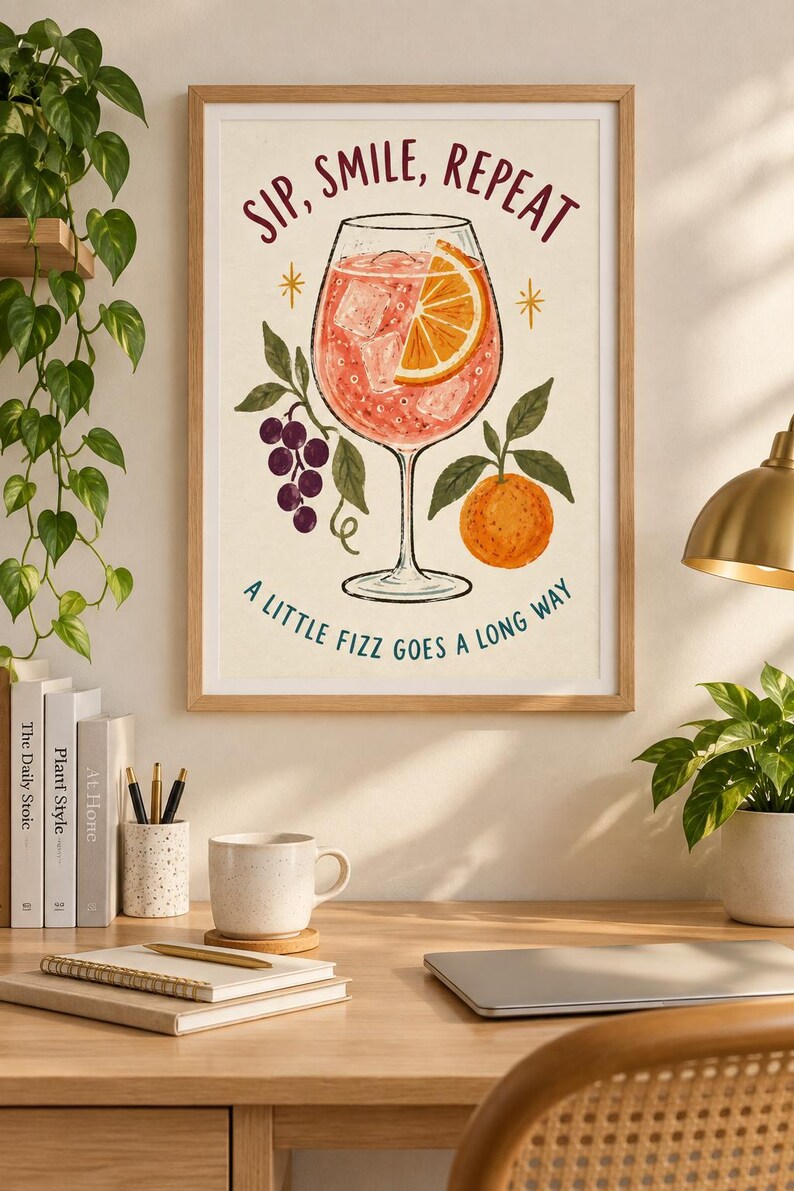 Op de afbeelding: Een ingelijste kunstprint met een cocktail in een glas op voet met een schijfje sinaasappel en ijsblokjes. De tekst luidt bovenaan SIP, SMILE, REPEAT en onderaan A LITTLE FIZZ GOES A LONG WAY, met illustraties van druiven en een sinaasappel.
