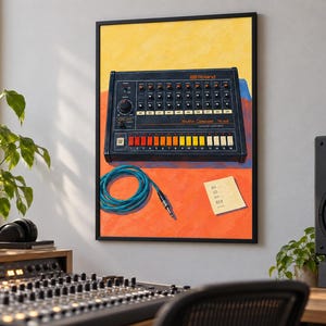 Op de afbeelding: Een ingelijste kunstdruk van een zwarte Roland TR-808 Rhythm Composer drumcomputer met oranje, gele en witte knoppen. Onder het apparaat liggen een opgerolde blauwe audiokabel en een klein geel briefje met tekst op een feloranje achtergrond.