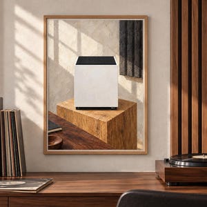 Op de afbeelding: Een ingelijste wandprint met een witte kubusvormige luidspreker met een zwart bovenpaneel, rustend op een getextureerd houten blok. Het kunstwerk hangt in een kamer met een houten platenspeler, een zwarte luidspreker en verticale houten wandlatten.