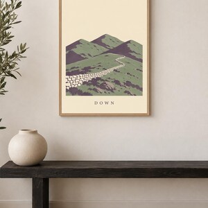 Pu&ograve; includere: Una stampa artistica incorniciata che presenta un'illustrazione paesaggistica stilizzata di montagne verdi e un muro di pietra che si snoda lungo un sentiero. La parola DOWN &egrave; stampata in lettere maiuscole scure al centro del fondo color crema.