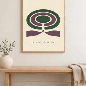 May include: A framed wall art print featuring a cream background with a centred graphic design of concentric dark green and purple oval shapes. Below the shapes, the text ROSCOMMON is printed in a simple, dark, sans-serif font.