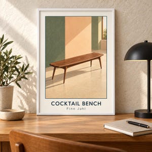 May include: A framed art print featuring a wooden bench with tapered legs set against a background of green and peach-colored walls. The text COCKTAIL BENCH and Finn Juhl is printed in dark blue at the bottom of the white-bordered poster.