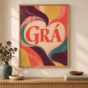 May include: A rectangular wall art print in a light wood frame featuring the word GR&Aacute; in large red letters centred within a cream-coloured heart shape, surrounded by abstract flowing shapes in shades of yellow, pink, teal, and deep purple.