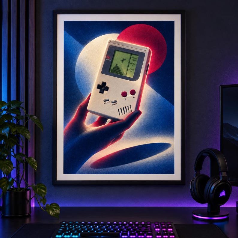 May include: A framed wall art print featuring a stylized illustration of a hand holding a vintage handheld gaming console. The console is light gray with a black directional pad, two red buttons, and a screen displaying a block-stacking game.