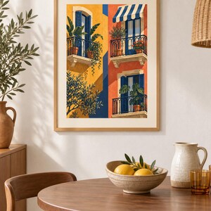 May include: A framed art print depicting a Mediterranean building facade with yellow and orange walls, blue shutters, and balconies filled with potted plants. The illustration features three levels of windows, a striped awning, and lush green foliage.