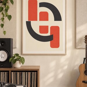 May include: A rectangular wall art print in a light wood frame featuring a graphic design of interlocking red and dark gray geometric shapes, including rounded rectangles and curved arcs, set against a solid off-white background.