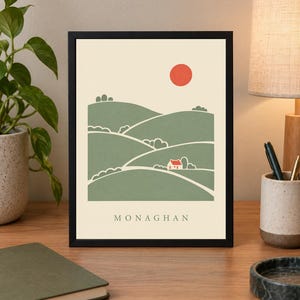 May include: A framed art print featuring a stylised landscape illustration of rolling green hills, a small white house with a red roof, and a solid red sun against a cream background, with the text MONAGHAN printed in black at the bottom.