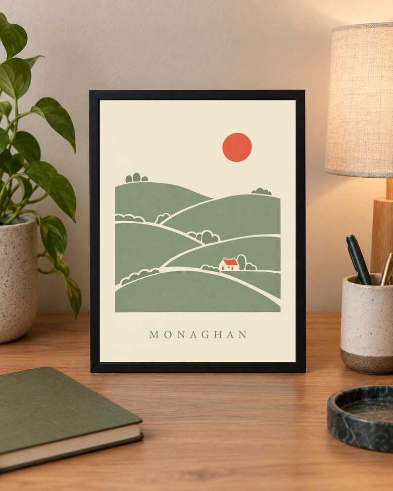 May include: A framed art print featuring a stylised landscape illustration of rolling green hills, a small white house with a red roof, and a solid red sun against a cream background, with the text MONAGHAN printed in black at the bottom.