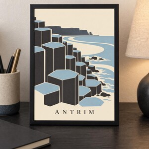 May include: A framed art print featuring a stylised illustration of the Giant's Causeway in Antrim. The image displays dark grey hexagonal basalt columns against a light blue sea and cream background, with the text ANTRIM printed at the bottom.