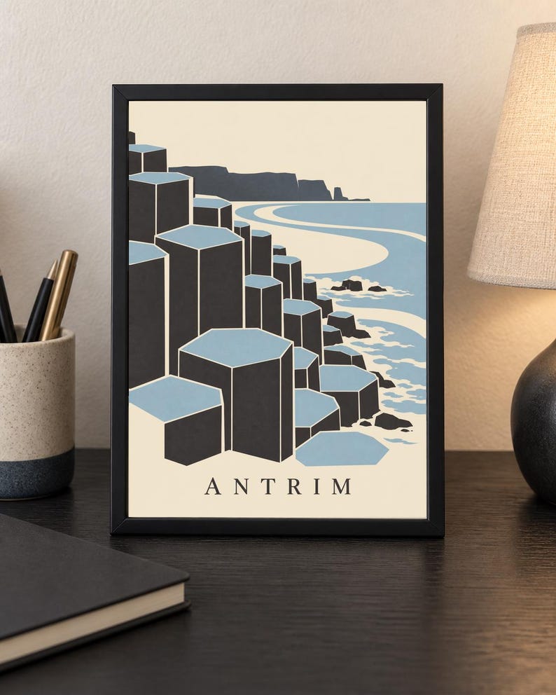 May include: A framed art print featuring a stylised illustration of the Giant's Causeway in Antrim. The image displays dark grey hexagonal basalt columns against a light blue sea and cream background, with the text ANTRIM printed at the bottom.