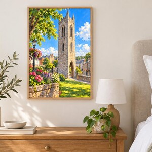 May include: A framed wall art print featuring a tall, weathered stone bell tower under a bright blue sky with white clouds. The scene includes a stone wall, vibrant pink and purple flowers, green trees, and a black lamp post in a sunny park.