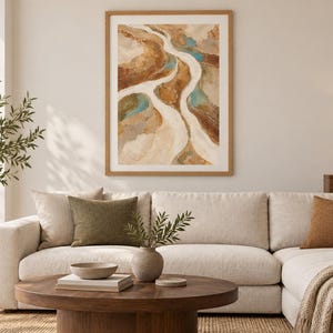 Pu&ograve; includere: Una stampa artistica da parete incorniciata che raffigura un dipinto di paesaggio astratto con forme bianche fluide simili a un fiume, terreno nei toni della terra marrone e marrone chiaro e piccole macchie di blu turchese brillante, in una cornice di legno chiaro.