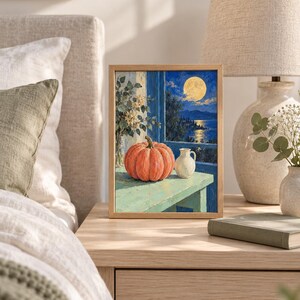 Pode incluir: Uma pintura retangular emoldurada retratando uma ab&oacute;bora laranja vibrante e um pequeno jarro branco sobre uma superf&iacute;cie verde-clara. A cena situa-se diante de uma janela com vista para um mar azul-escuro sob uma lua cheia brilhante.
