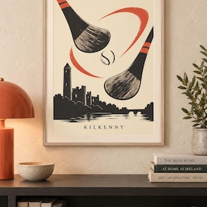 Peut inclure: Une affiche encadr&eacute;e repr&eacute;sentant une illustration stylis&eacute;e de deux crosses de hurling et une balle au-dessus de la silhouette du ch&acirc;teau et de la tour ronde de Kilkenny. Le design utilise un fond cr&egrave;me avec des accents noirs et orange et le texte KILKENNY.
