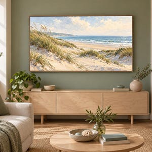 May include: A rectangular landscape painting in a thin wooden frame depicting a coastal scene with sand dunes, tall beach grass, and a blue ocean under a partly cloudy sky, mounted on a sage green wall above a light wood media console.