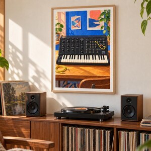 May include: A framed wall art print featuring a black Korg MS-20 analog synthesizer with a yellow patch cable, set against a vibrant blue wall with two small landscape paintings and a potted plant on a wooden desk surface.