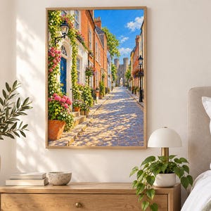 May include: A framed wall art print depicting a cobblestone street lined with colorful brick buildings, vibrant pink climbing flowers, and green foliage. The scene features a blue door, a black street lamp, and a stone castle gate in the distance.