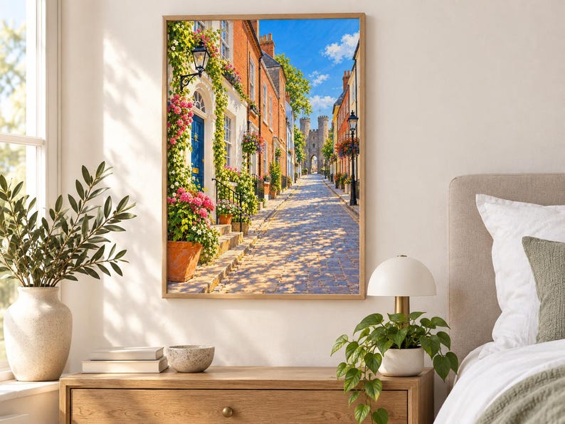 May include: A framed wall art print depicting a cobblestone street lined with colorful brick buildings, vibrant pink climbing flowers, and green foliage. The scene features a blue door, a black street lamp, and a stone castle gate in the distance.
