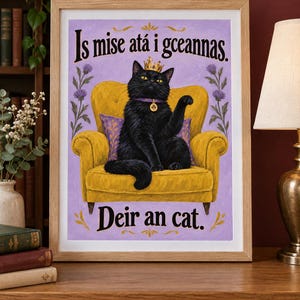 May include: A framed art print featuring a black cat wearing a gold crown and a purple collar with a clover charm, sitting on a yellow armchair against a light purple background. The text reads Is mise at&aacute; i gceannas. Deir an cat.