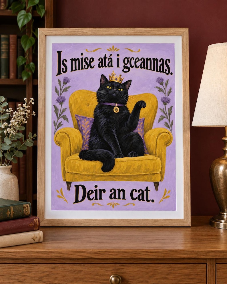 May include: A framed art print featuring a black cat wearing a gold crown and a purple collar with a clover charm, sitting on a yellow armchair against a light purple background. The text reads Is mise at&aacute; i gceannas. Deir an cat.