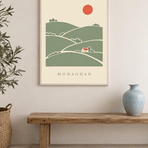 May include: A rectangular wall art print in a light frame featuring a stylised landscape illustration of green rolling hills, a small white house with a red roof, and a solid red sun against a cream background with the text MONAGHAN below.