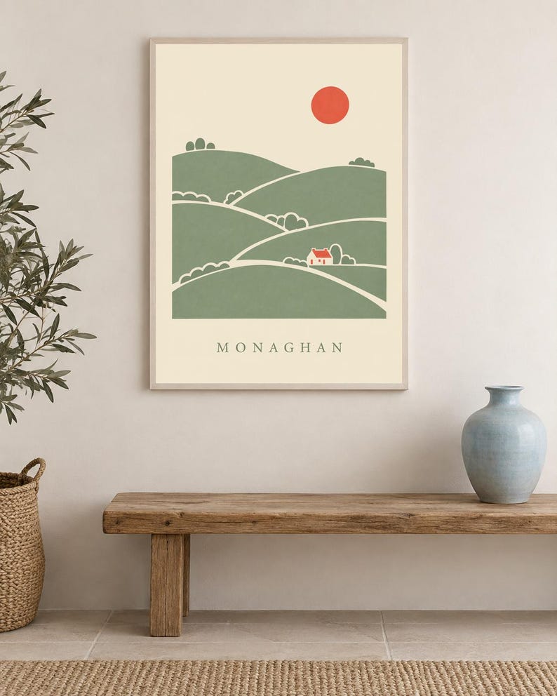 May include: A rectangular wall art print in a light frame featuring a stylised landscape illustration of green rolling hills, a small white house with a red roof, and a solid red sun against a cream background with the text MONAGHAN below.