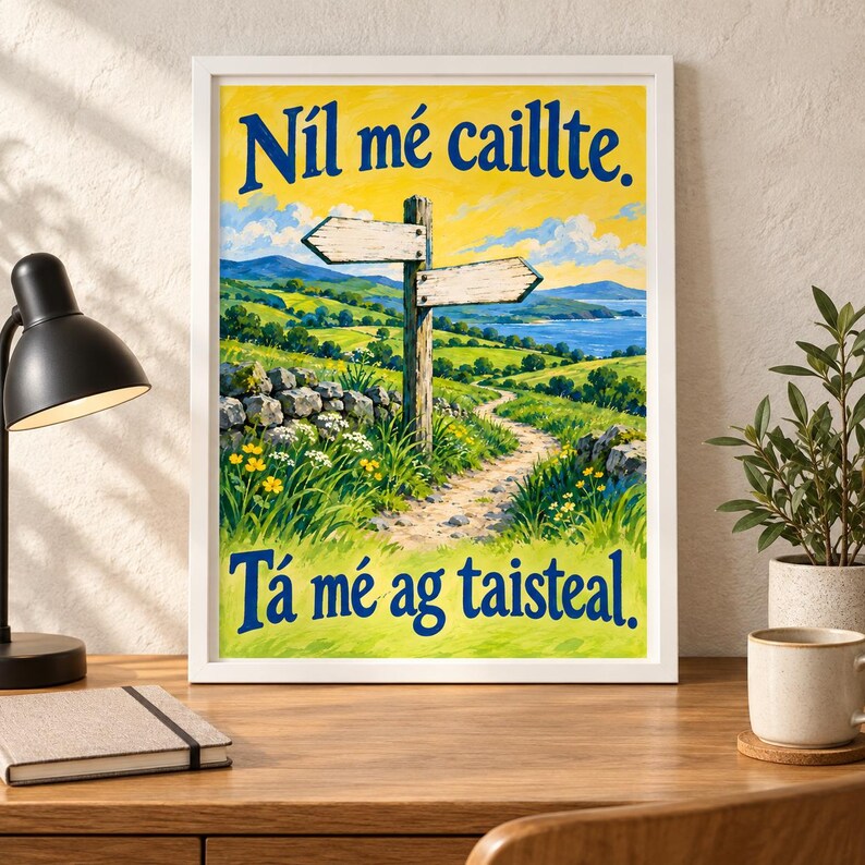 Op de afbeelding: Een ingelijste kunstdruk met een landschapsillustratie van een zandpad, groene heuvels en een houten wegwijzer. De tekst "N&iacute;l m&eacute; caillte. T&aacute; m&eacute; ag taisteal." staat in blauwe letters gedrukt tegen een gele lucht op de achtergrond.
