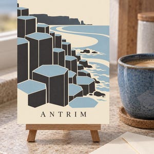 May include: A rectangular print on a small wooden easel featuring a stylised illustration of the Giant's Causeway in Antrim. The artwork displays dark grey hexagonal basalt columns against a light blue sea and cream background with the text ANTRIM.
