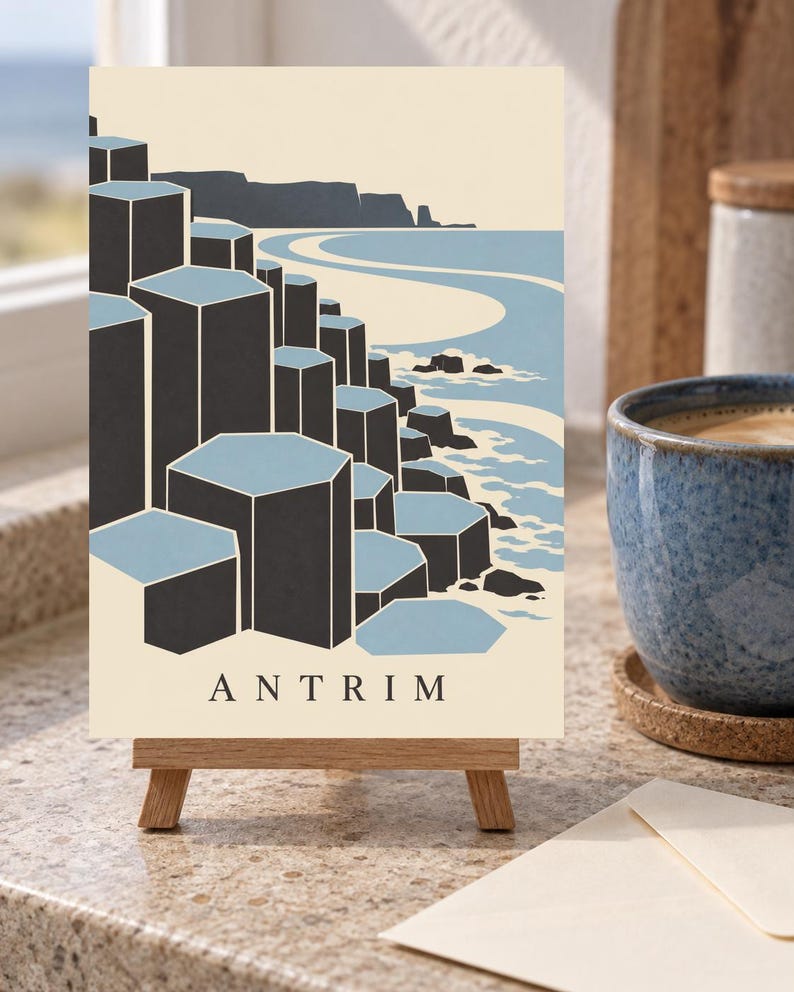 May include: A rectangular print on a small wooden easel featuring a stylised illustration of the Giant's Causeway in Antrim. The artwork displays dark grey hexagonal basalt columns against a light blue sea and cream background with the text ANTRIM.