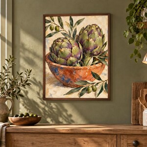 May include: A framed wall art print featuring a painting of two green and purple artichokes resting in a rustic, textured orange bowl. The artwork includes sprigs of green olive leaves and small olives, rendered in a painterly style with visible brushstrokes.