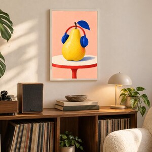 May include: A framed wall art print featuring a yellow pear wearing blue headphones with a red headband, set against a peach background. The pear sits on a white circular table with a red base, depicted in a flat, graphic illustration style.