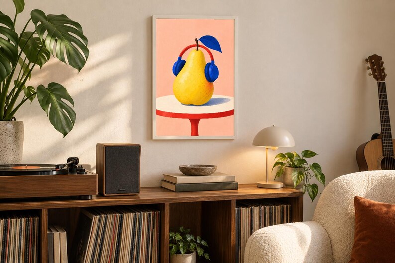 May include: A framed wall art print featuring a yellow pear wearing blue headphones with a red headband, set against a peach background. The pear sits on a white circular table with a red base, depicted in a flat, graphic illustration style.