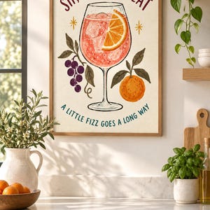 Op de afbeelding: Een ingelijste wandprint met een cocktail in een glas op voet met een schijfje sinaasappel en ijsblokjes. De tekst luidt bovenaan SIP, SMILE, REPEAT en onderaan A LITTLE FIZZ GOES A LONG WAY, met illustraties van druiven en een sinaasappel.