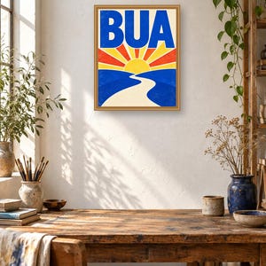 May include: A framed rectangular wall art print featuring the word BUA in bold blue letters above a stylized yellow sun with red and yellow rays. Below the sun, a winding white path leads through blue hills, all set against a cream background.