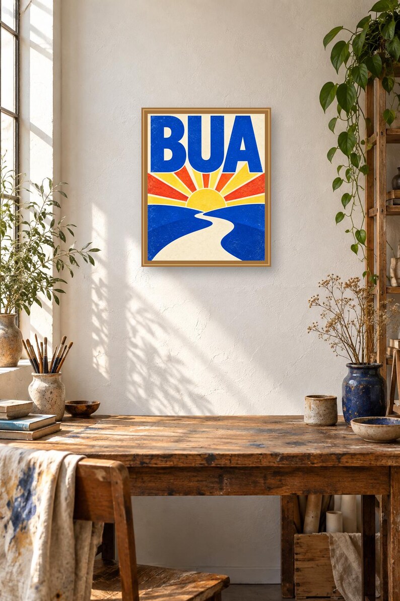 May include: A framed rectangular wall art print featuring the word BUA in bold blue letters above a stylized yellow sun with red and yellow rays. Below the sun, a winding white path leads through blue hills, all set against a cream background.