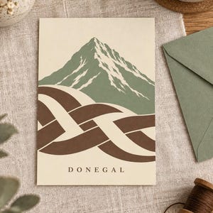 K&ouml;nnte beinhalten: Eine rechteckige Gru&szlig;karte mit einer grafischen Illustration eines gr&uuml;nen Berges mit wei&szlig;en Schneegipfeln &uuml;ber einem braun-cremefarbenen, ineinander verschlungenen Knotenmuster. Das Wort DONEGAL ist in schwarzen Gro&szlig;buchstaben unten aufgedruckt.