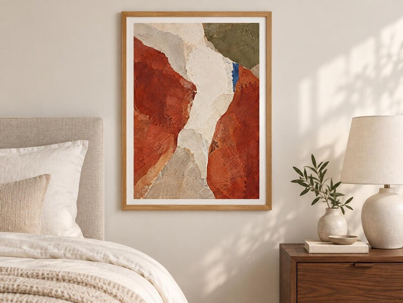 May include: A framed abstract wall art print featuring a textured composition of deep terracotta red, muted olive green, off-white, and light gray shapes. The artwork is enclosed in a simple light-toned wooden frame with a white mat border.