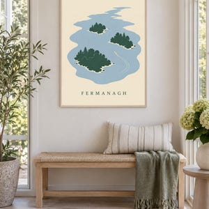 May include: A rectangular wall art print featuring a light blue lake with three dark green islands covered in trees. A small white boat leaves a wake on the water. The background is cream-colored, and the word FERMANAGH is printed in black.