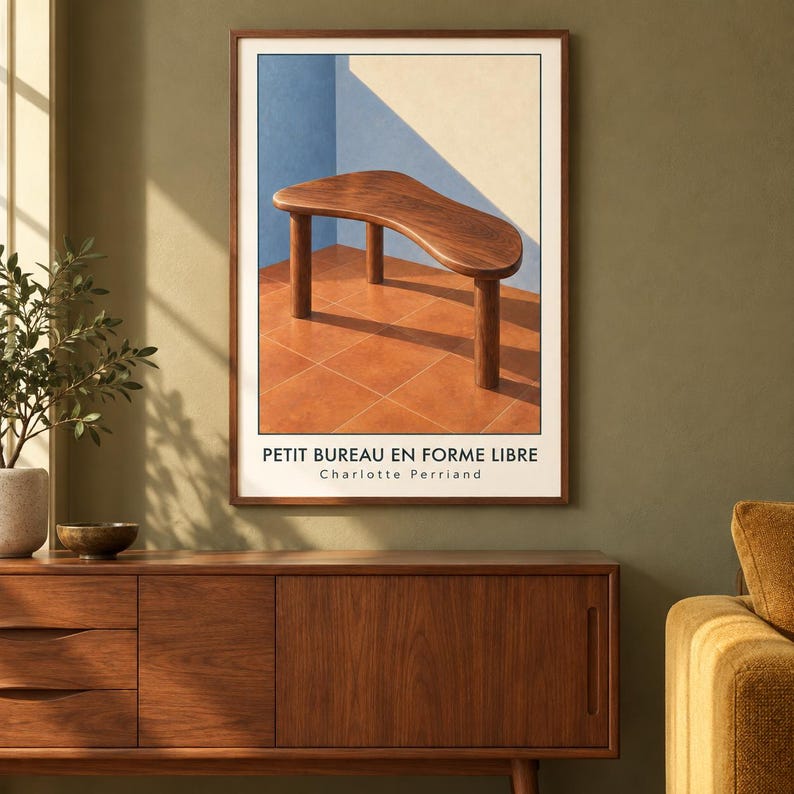May include: A framed art print featuring a dark wood desk with a free-form curved top and three cylindrical legs, set against a blue and cream wall on a terracotta tiled floor. The text reads PETIT BUREAU EN FORME LIBRE Charlotte Perriand.