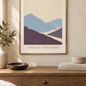 May include: A framed wall art print featuring a landscape illustration of layered mountains in shades of blue and deep purple against a cream background, with the text COOLEY STILLNESS printed in black capital letters at the bottom of the image.