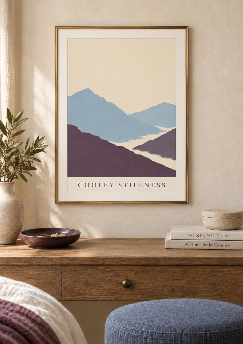 May include: A framed wall art print featuring a landscape illustration of layered mountains in shades of blue and deep purple against a cream background, with the text COOLEY STILLNESS printed in black capital letters at the bottom of the image.