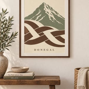 K&ouml;nnte beinhalten: Ein gerahmter Wandkunstdruck mit einem stilisierten Berggipfel in gedecktem Gr&uuml;n und Wei&szlig;, platziert &uuml;ber einem abstrakten braun-cremefarbenen keltischen Knotenmuster. Das Wort DONEGAL ist in schwarzen Gro&szlig;buchstaben auf dem cremefarbenen Hintergrund gedruckt.